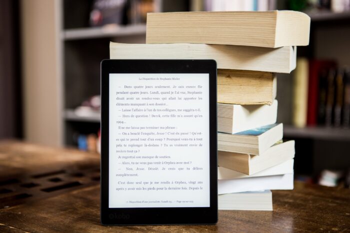 An Amazon Kindle e-reader positioned in the foreground with a blurred background of a bookshelf filled with various books, illustrating the bridge between traditional reading and modern digital technology.