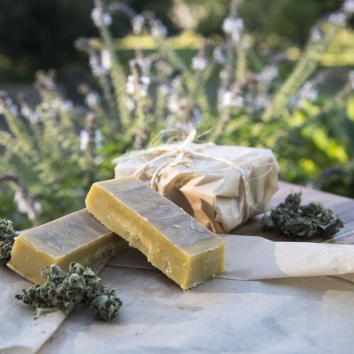 Blocks of cannabis butter on a wooden cutting board, surrounded by cannabis buds and wrapped in parchment paper, with a natural outdoor background